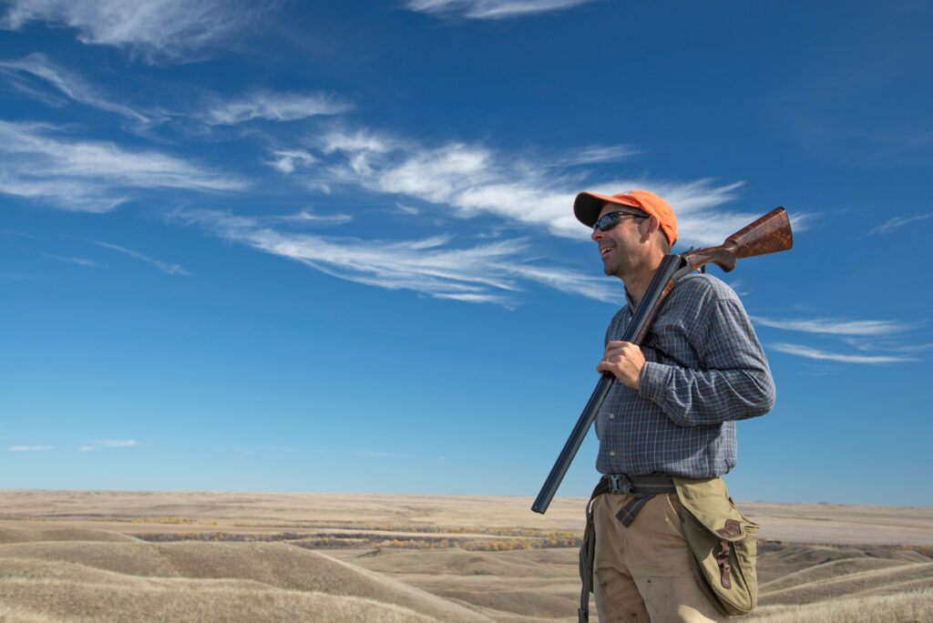 An upland bird hunter in front of a blue sky in an open western prairie holding a shotgun over his shoulder.