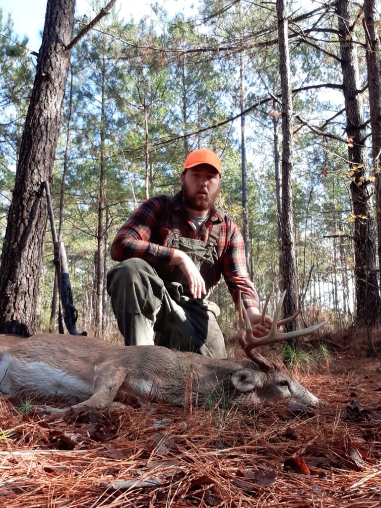 A deer hunter in a pine forest holding up a very large, mature, old whitetail buck trophy deer.