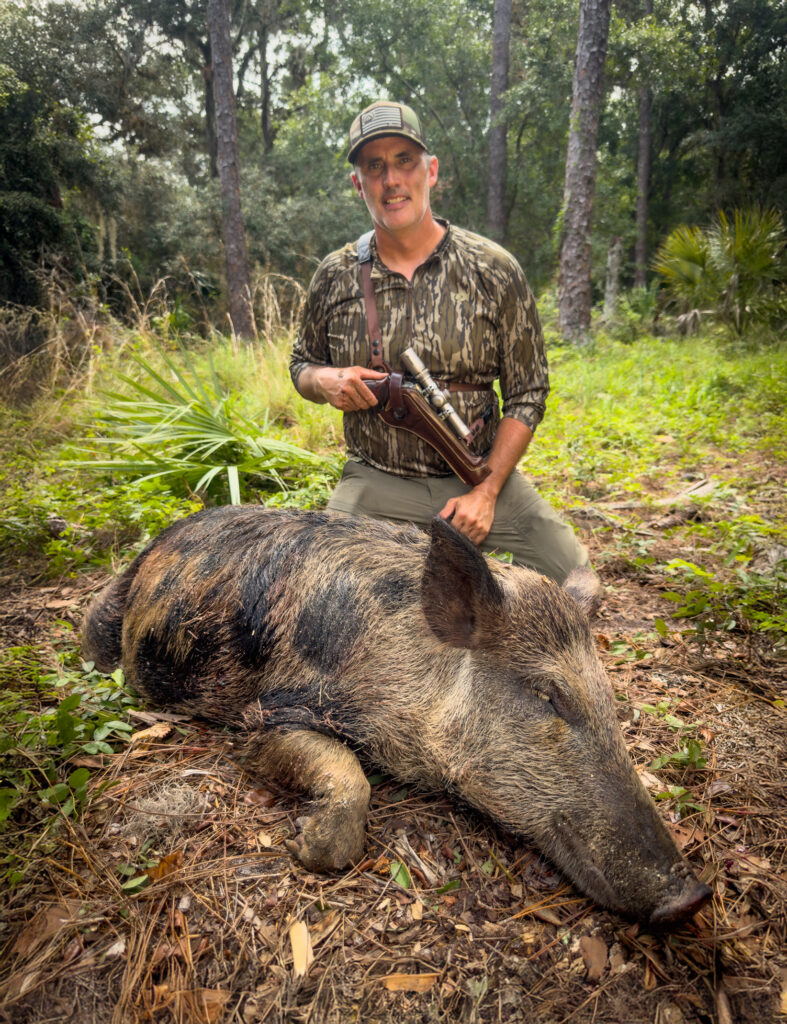A hunter holding a scoped revolver handgun with a dead hog after a successful hunt.