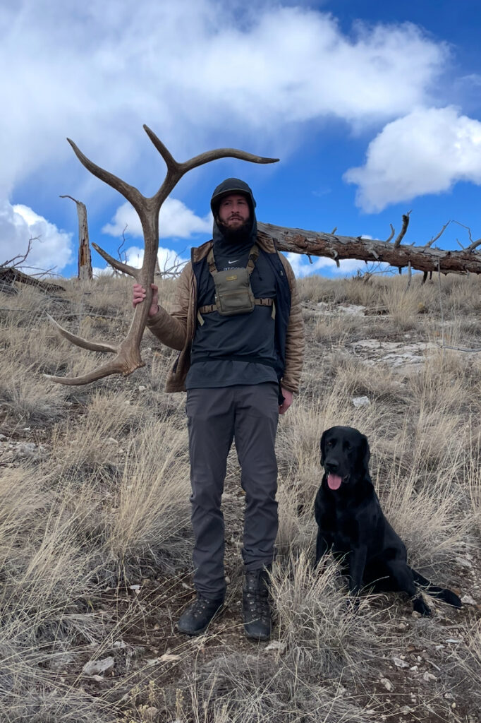 A man with a black Lab holds an elk antler while shed hunting.